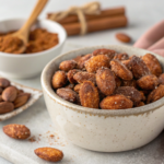 A bowl of homemade cinnamon almonds, coated in a light dusting of cinnamon and coconut sugar, served on a wooden table with a cinnamon stick beside them.